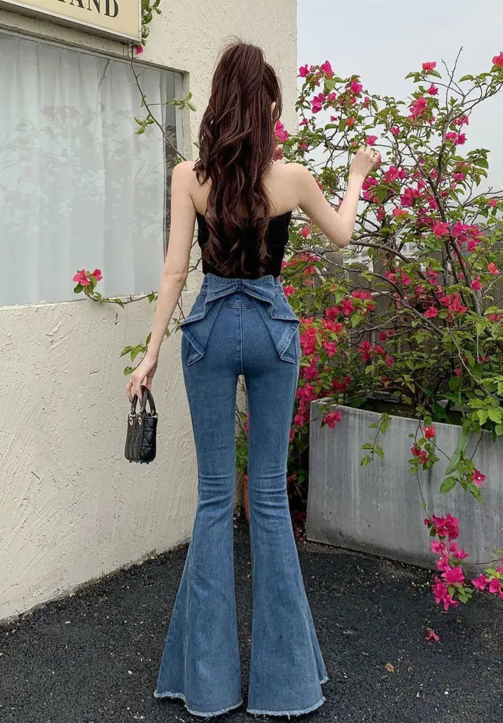 Model standing in front of flowers, showing the back view of the Dark Blue Backstage Bow Flare Jeans, showcasing the bow detail.