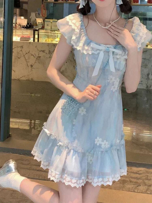Model wearing the mini Ethereal Flower Princess Dress in soft blue with ruffle trim and daisy embroidery, standing near a glass window.