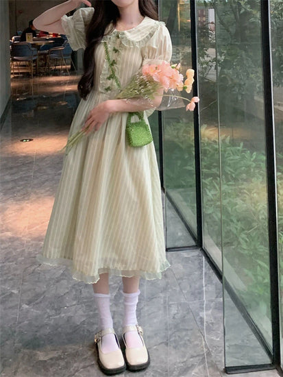 Model in midi-length Pistachio Ribbon Striped Dress holding peach flowers, standing by a glass wall.