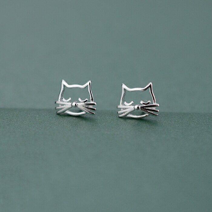 Silver Kitty Stud Earrings shown together from the front on a muted green background.
