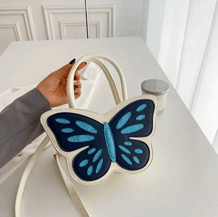 Model holding Sparkling Butterfly Handbag in white and blue by the handle on a table