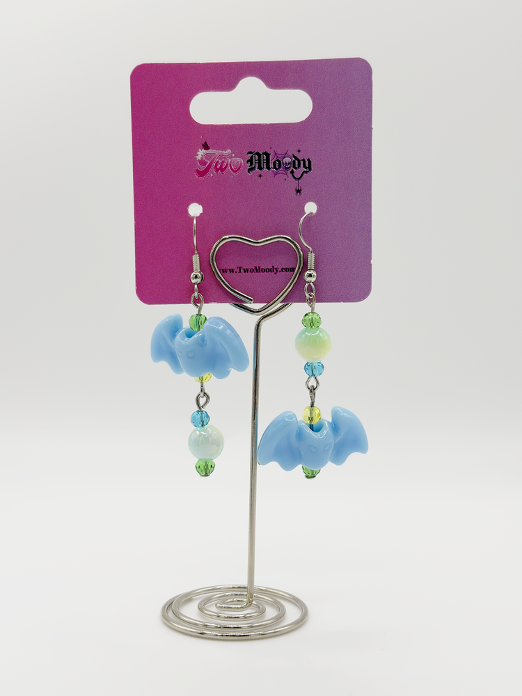 Two Moody Originals Candy Bite Bat Earrings featuring pastel blue bat charms with green, blue, and yellow glass and acrylic beads in mismatched opposite designs hanging on a Two Moody jewelry display.