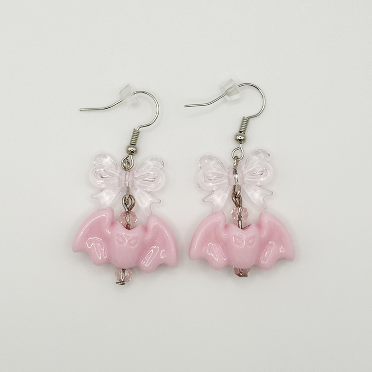 Two Moody Originals Candy Bite Bat Earrings with pastel pink bat charms, transparent pink acrylic bows, and pink glass bead accents.
