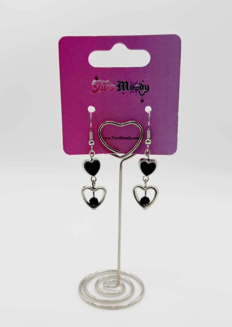 Two Moody Originals: Lovesick Charm Earrings with black crystal heart accents and glossy black enamel heart charms on a Two Moody jewelry display.