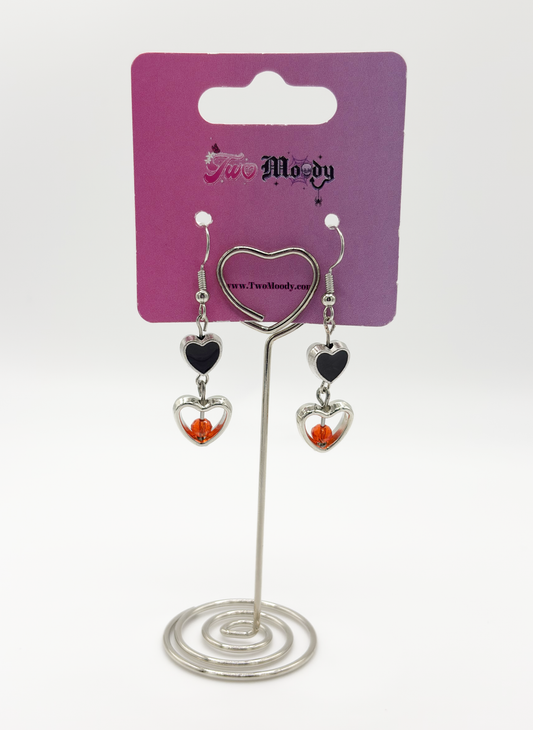 Two Moody Originals: Lovesick Charm Earrings with red-orange crystal heart accents and silver dangling heart details on a Two Moody jewelry display.