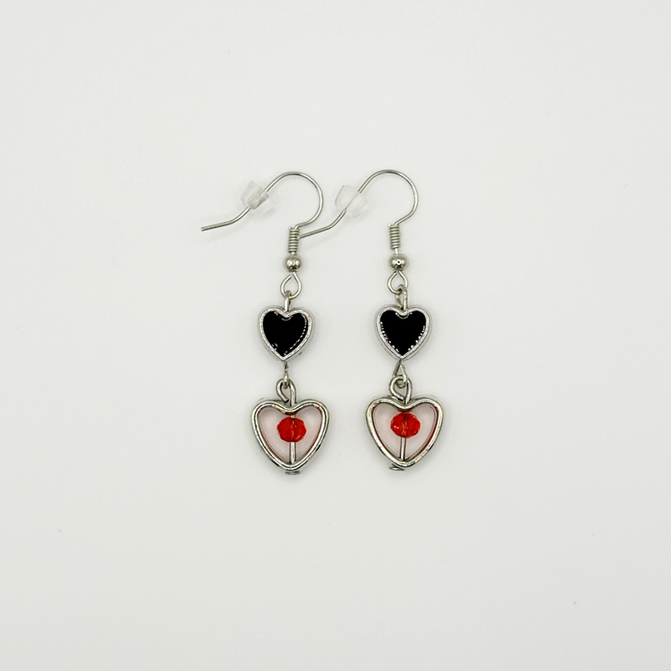 Two Moody Originals: Lovesick Charm Earrings with black enamel heart charms, silver open heart drops, and red-orange crystal bead accents.