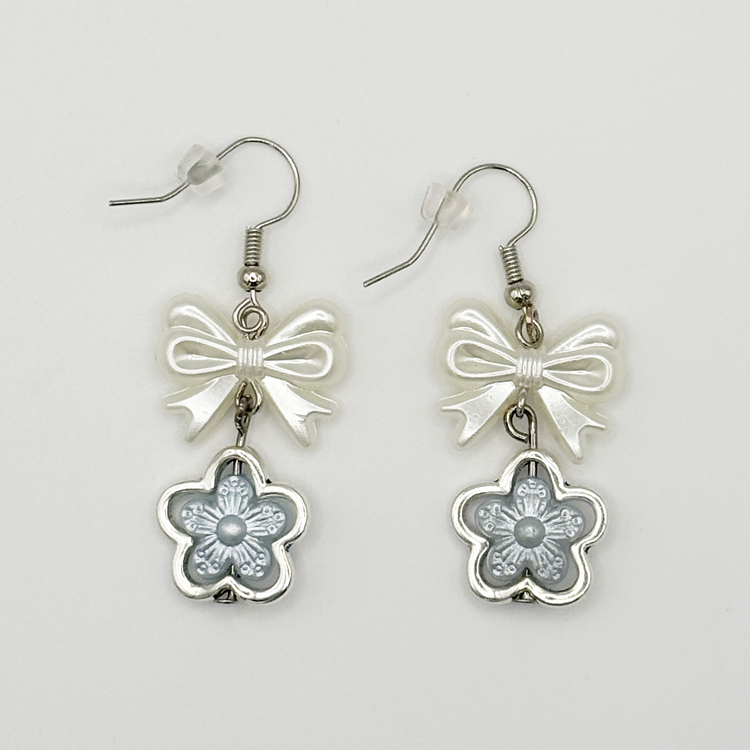 Two Moody Originals: Soft Bloom Bow Earrings with glossy white bow charms and silver and icy blue floral pendants on silver hooks.