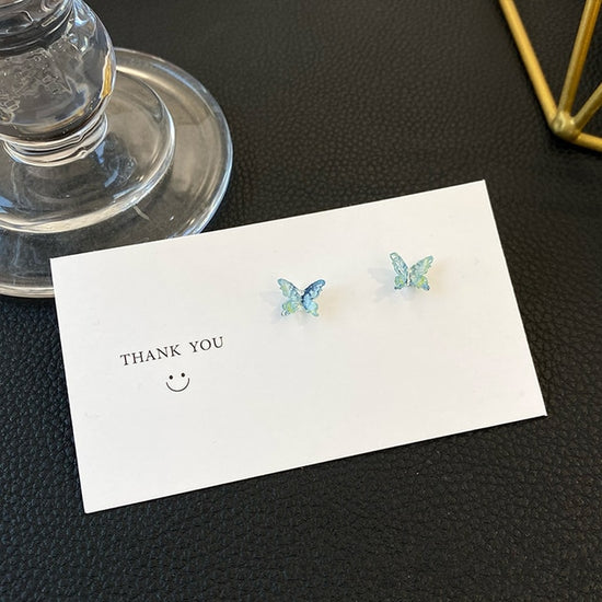 A pair of Dreamlit Butterfly Stud Earrings in a cool blue-green shade styled on a simple card.