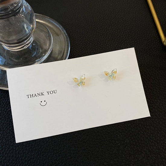 A soft yellowish-blue version of the Dreamlit Butterfly Stud Earrings set against a white card.