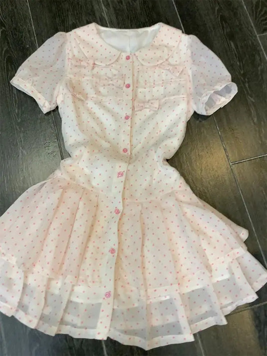 The front of the Dollhouse Dream Dress, a short-sleeve white dress with a light pink polka dot pattern, featuring a button-up front, bows, and a tiered pleated flared skirt, laid flat on a dark wood floor.