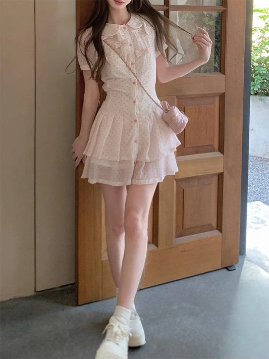 A model wearing the Dollhouse Dream Dress, a short-sleeve white dress with pink polka dots and a tiered pleated skirt, standing in front of a wooden door and holding a matching pink crossbody bag.