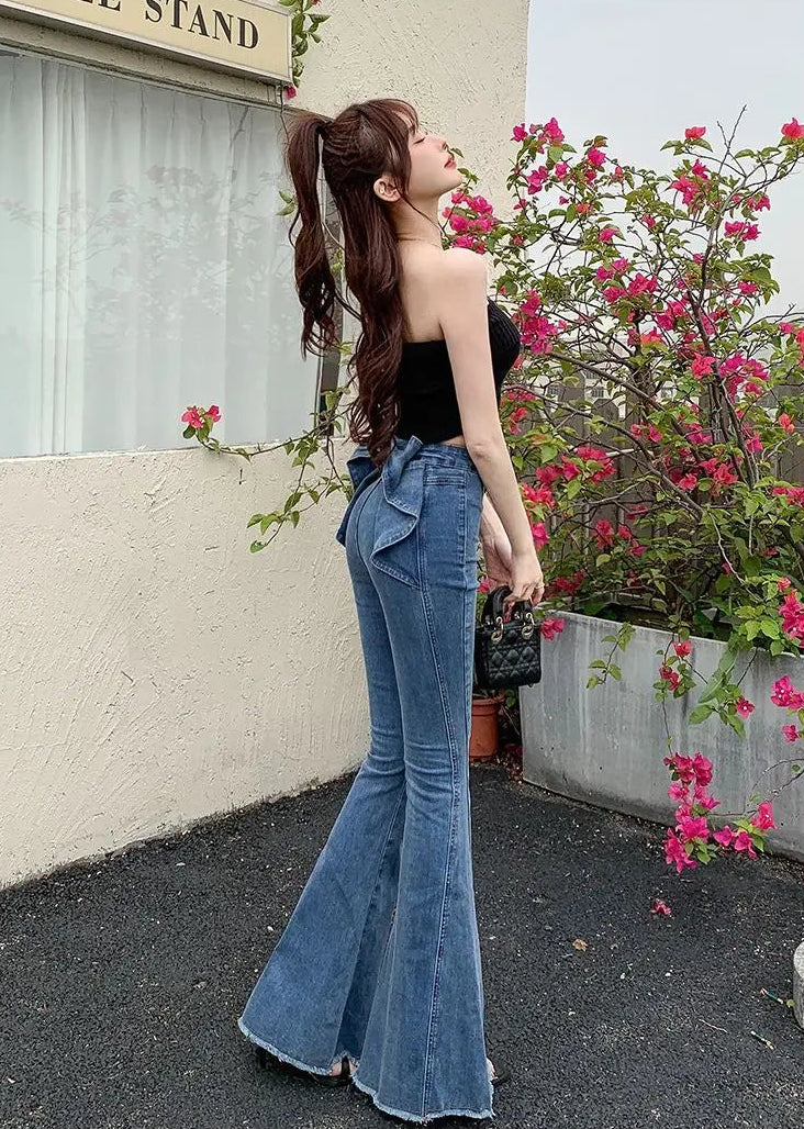 Model standing in front of flowers at an angle, showing the back view of the Dark Blue Backstage Bow Flare Jeans, showcasing the side of the bow detail.