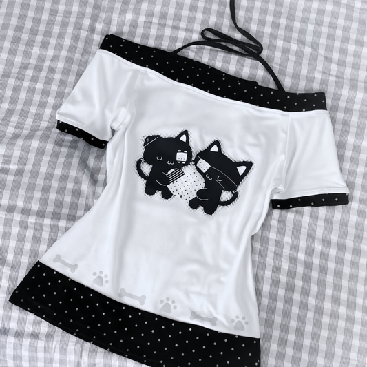Flat lay of the Bandaged Kitties Off-Shoulder Top showcasing the adjustable halter strap, off-shoulder neckline, bandaged kitty graphic, contrasting polka dot trim, and decorative paw print and bone accents along the hem.
