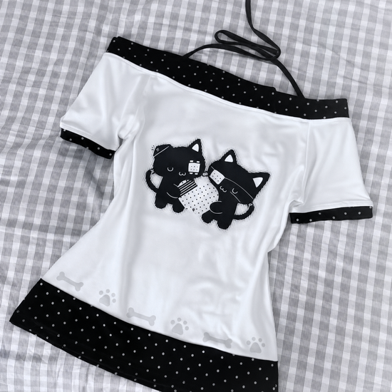 Flat lay of the Bandaged Kitties Off-Shoulder Top showcasing the adjustable halter strap, off-shoulder neckline, bandaged kitty graphic, contrasting polka dot trim, and decorative paw print and bone accents along the hem.