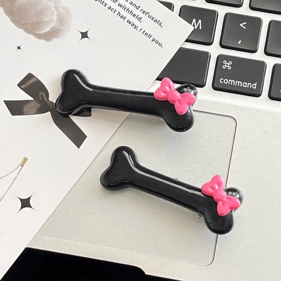 Pair of black Bowtiful Bones Hair Clips, featuring glossy bone-shaped designs with pink bow accents.