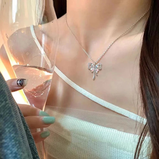 A model wearing the Celestial Ribbon Bow Necklace, a silver bow pendant necklace, while holding a glass in warm sunlight.