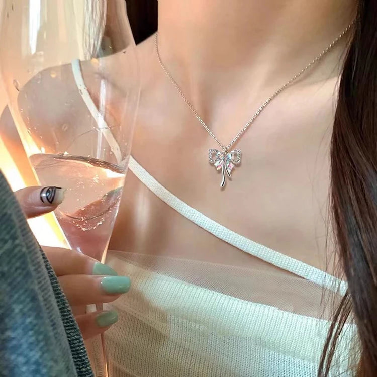A model wearing the Celestial Ribbon Bow Necklace, a silver bow pendant necklace, while holding a glass in warm sunlight.