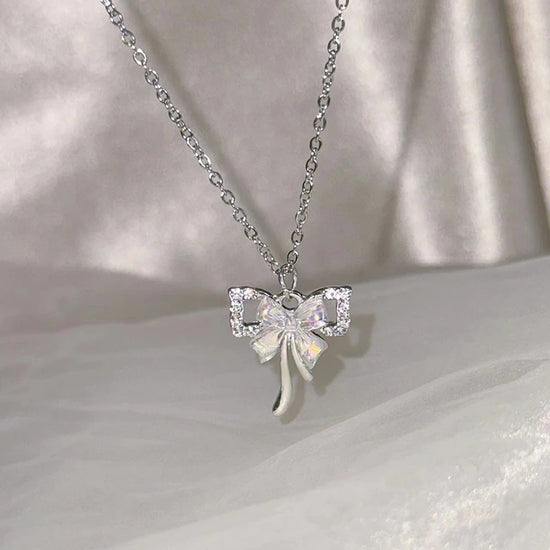 The Celestial Ribbon Bow Necklace, a silver crystal bow pendant necklace, displayed above soft white satin.