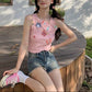 Model wearing the dusty pink Charm Collector Tank with denim shorts in a side seated pose on a wooden deck.