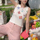Model wearing the white Charm Collector Tank with pink pants beside a colorful outdoor fruit stand.