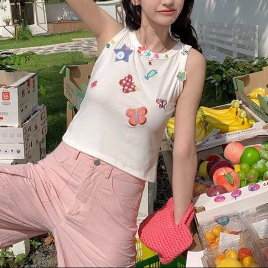 Model wearing the white Charm Collector Tank with pink pants beside a colorful outdoor fruit stand.