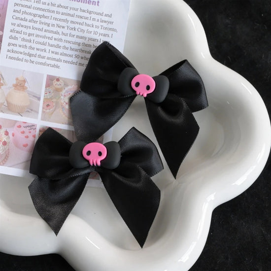 Creep It Cute Hair Bows — Pair of black satin hair bows with pink skull centers on a white flower-shaped tray.
