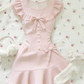 The Dotted Darling Dress, a pink polka-dot mini dress, lies flat on a floral bedding with bow and ruffle details.