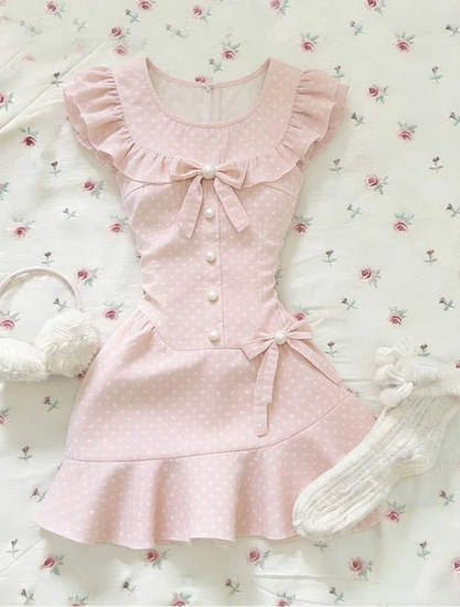 The Dotted Darling Dress, a pink polka-dot mini dress, lies flat on a floral bedding with bow and ruffle details.