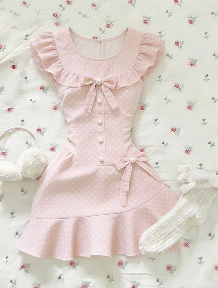 The Dotted Darling Dress, a pink polka-dot mini dress, lies flat on a floral bedding with bow and ruffle details.