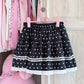 Dotted Doll Lace Skort in black with white polka dots, gathered tiers, and white lace trim displayed on a pink hanger.