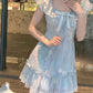Model wearing the mini Ethereal Flower Princess Dress in soft blue with ruffle trim and daisy embroidery, standing near a glass window.