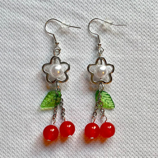 Close-up of Fairy Cherry Pearl Earrings with pearl bead and red cherry charm on a white textured background