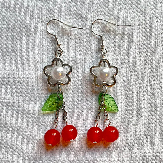 Close-up of Fairy Cherry Pearl Earrings with pearl bead and red cherry charm on a white textured background