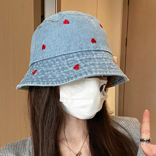 A model wearing the Love Stitch Denim Bucket Hat in light blue, showing the soft denim fabric and delicate red heart embroidery.