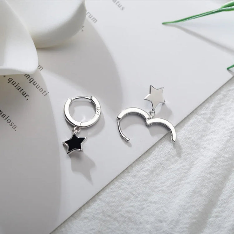The Midnight Star Drop Earrings, black star-shaped hoop earrings, displayed on a decorated table, with one earring open and shown from the front and the other open and shown from the back.