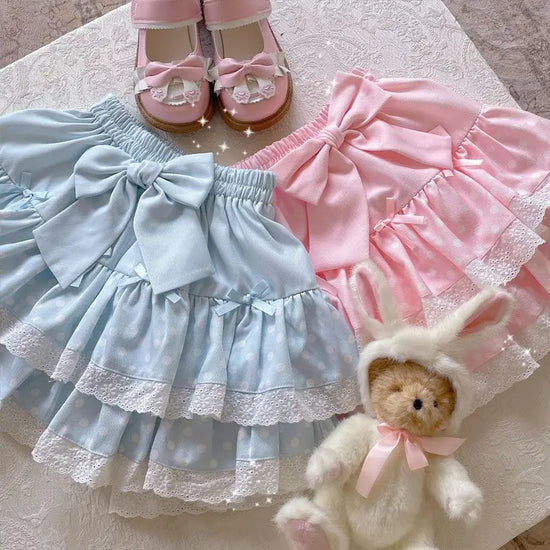 Pink and blue versions of the Polka Dot Sweetie Tiered Skirt displayed side by side with lace ruffles, ribbon accents, and removable oversized bow pins.