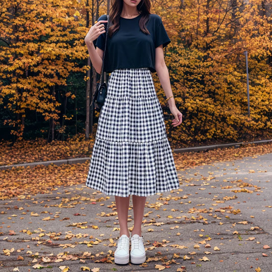 A model wearing the Quiet Charm Gingham Midi Skirt, a black and white gingham midi skirt, with a black tee and white sneakers on an autumn tree-lined path.