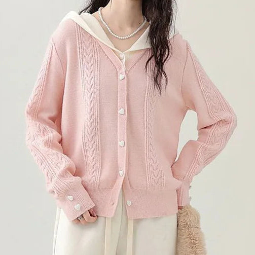 Model wearing the Sweetheart Cable Knit Cardigan in light pink with a cream hood, posing with one hand on her hip to show the heart-shaped buttons and cable knit texture.
