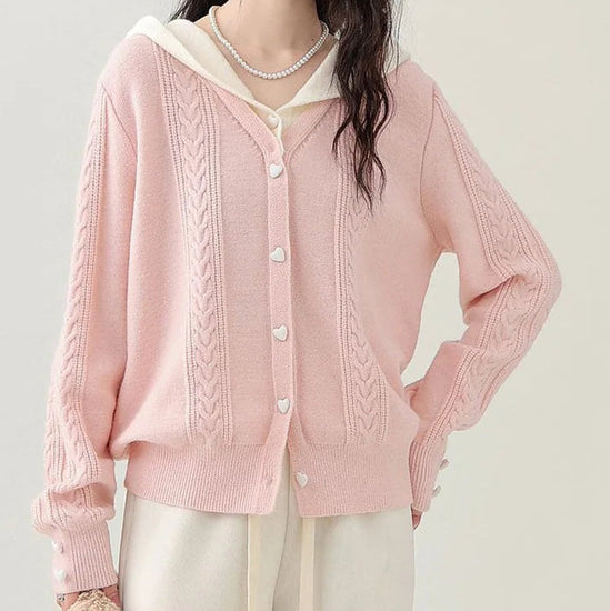 Model wearing the Sweetheart Cable Knit Cardigan in light pink with cream hood, showing the heart-shaped buttons and cable knit detailing.