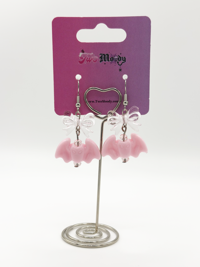 Two Moody Originals Candy Bite Bat Earrings with pastel pink bat charms, transparent pink acrylic bows, and pink glass bead accents on a Two Moody jewelry display.