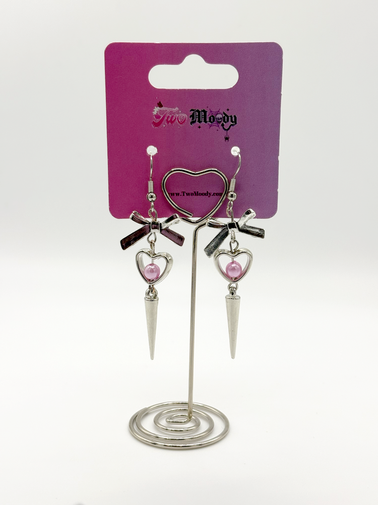 Two Moody Originals: Coquette Dagger Earrings displayed on branded packaging with silver heart charms featuring pink faux pearl accents, silver bows, and dagger drops.