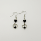 Two Moody Originals: Lovesick Charm Earrings with glossy black heart charms, silver open heart drops, and black crystal bead accents.