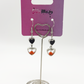 Two Moody Originals: Lovesick Charm Earrings with red-orange crystal heart accents and silver dangling heart details on a Two Moody jewelry display.