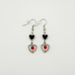 Two Moody Originals: Lovesick Charm Earrings with black enamel heart charms, silver open heart drops, and red-orange crystal bead accents.