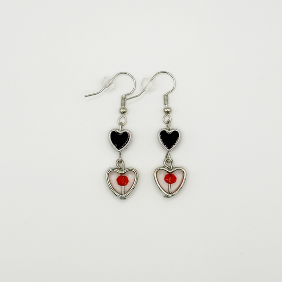 Two Moody Originals: Lovesick Charm Earrings with black enamel heart charms, silver open heart drops, and red-orange crystal bead accents.