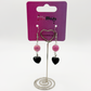 The Moody Heartbreaker Earrings displayed on a card, highlighting the pink bead and black heart charm details.