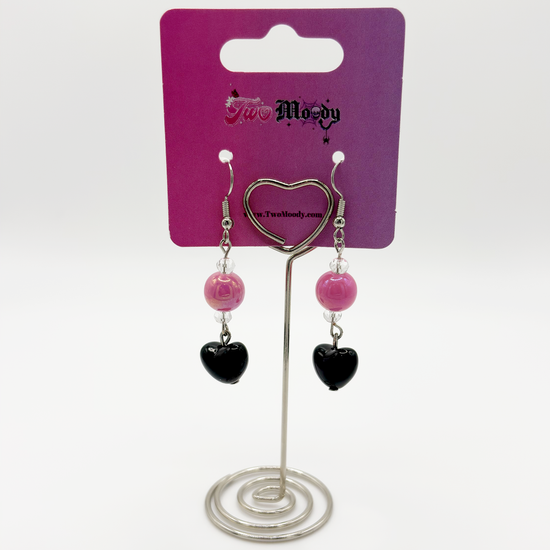 The Moody Heartbreaker Earrings displayed on a card, highlighting the pink bead and black heart charm details.