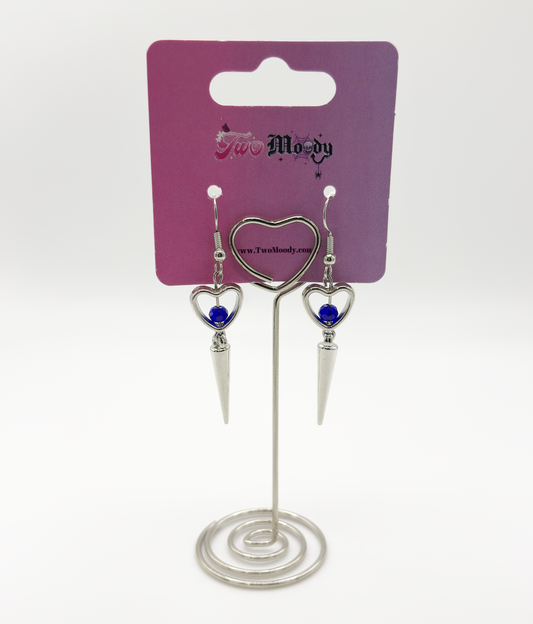 Two Moody Originals: Pierced Heart Dagger Earrings displayed on branded packaging with silver open heart charms, blue crystal accents, and dangling dagger details.