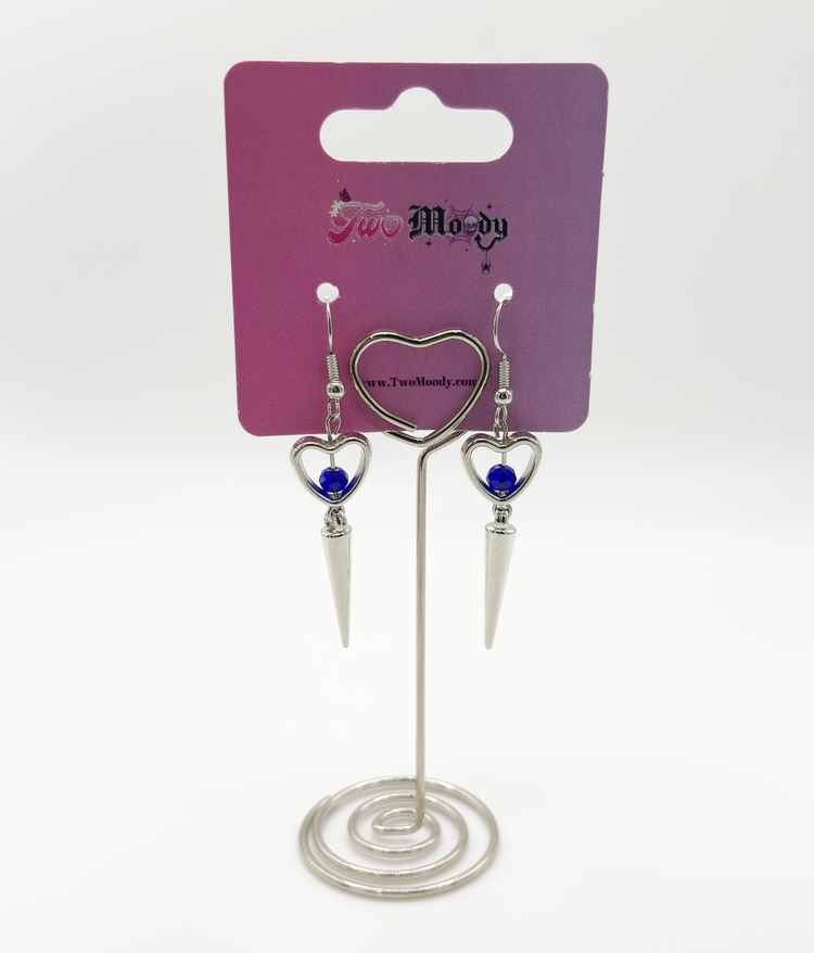 Two Moody Originals: Pierced Heart Dagger Earrings displayed on branded packaging with silver open heart charms, blue crystal accents, and dangling dagger details.