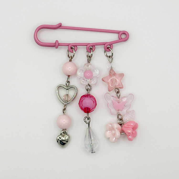 Two Moody Originals: Pink Daydream Pin featuring dangling pastel charms, silver heart details, butterfly accents, and crystal beads.